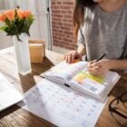 A imagem mostra uma mulher organizando sua agenda em uma mesa de madeira. Ela está escrevendo em um planner colorido, cercada por um calendário e um laptop. Um vaso com flores laranjas decora o ambiente, transmitindo uma sensação de produtividade e organização. A luz natural ilumina o espaço, criando um ambiente acolhedor e inspirador.