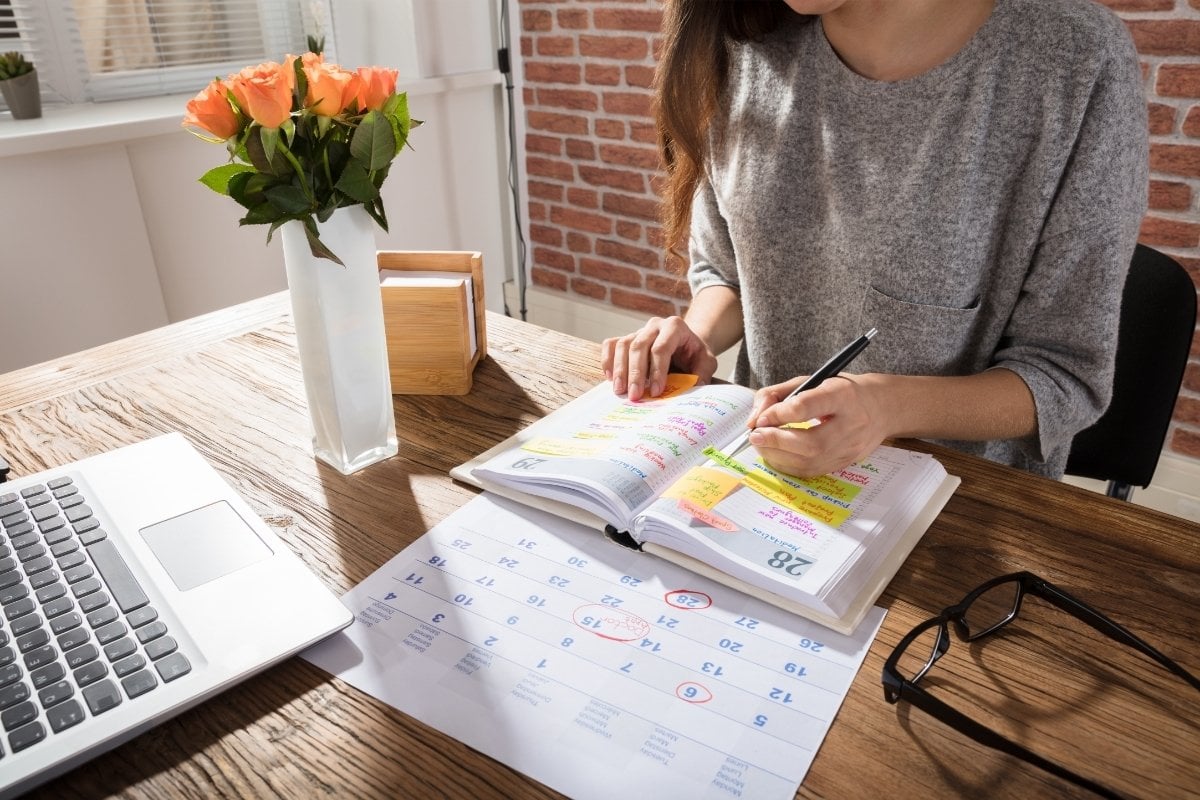 A imagem mostra uma mulher organizando sua agenda em uma mesa de madeira. Ela está escrevendo em um planner colorido, cercada por um calendário e um laptop. Um vaso com flores laranjas decora o ambiente, transmitindo uma sensação de produtividade e organização. A luz natural ilumina o espaço, criando um ambiente acolhedor e inspirador.