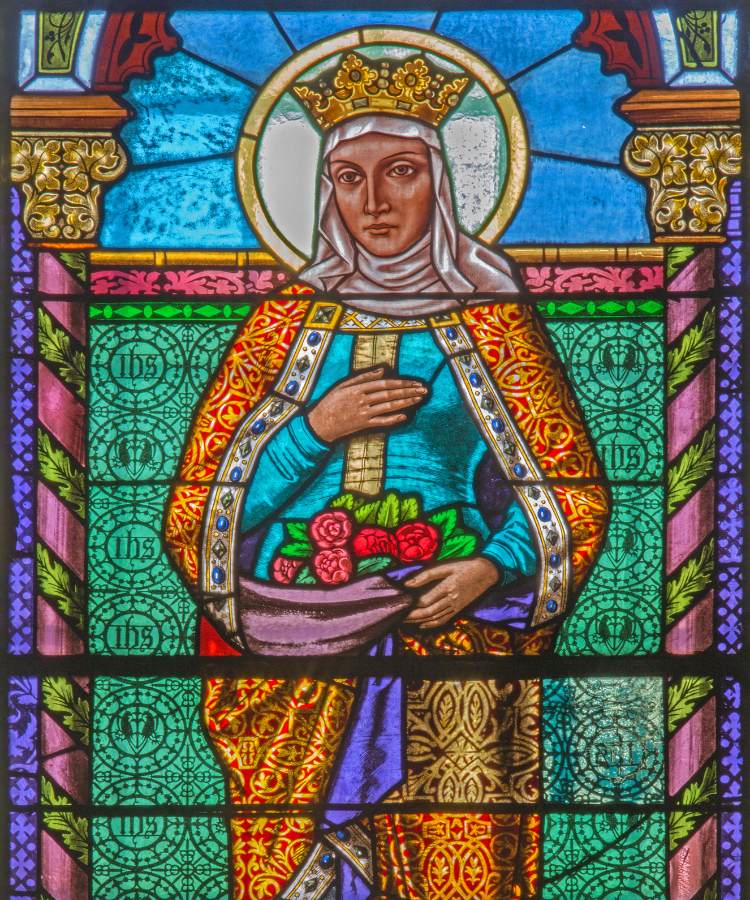 O vitral representa Santa Isabel da Hungria com uma coroa dourada e um manto ricamente decorado. Ela segura rosas em suas mãos, simbolizando seu milagre. O fundo é azul e verde, com detalhes ornamentais, destacando sua figura serena e majestosa. O halo dourado ao redor da cabeça enfatiza sua santidade.