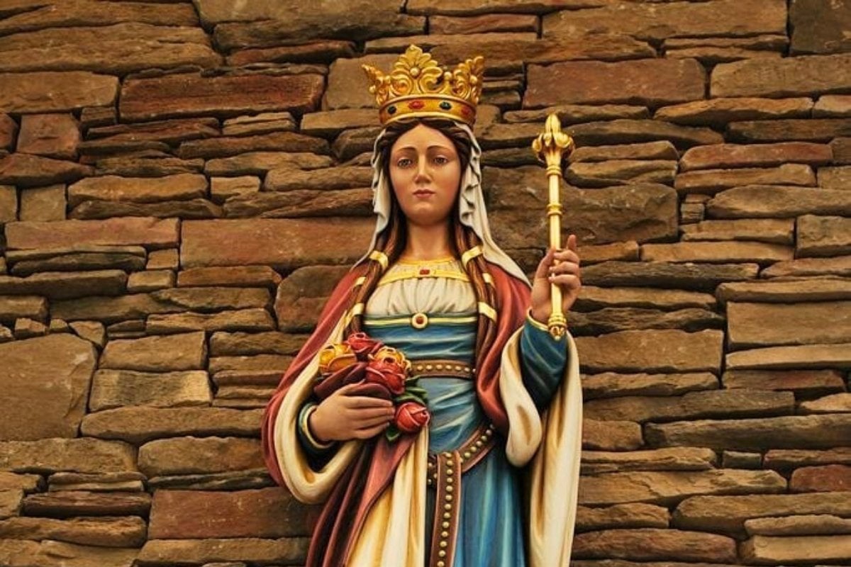 A imagem retrata uma estátua de Santa Isabel da Hungria, vestida com trajes reais. Ela usa uma coroa dourada adornada com pedras preciosas e segura um cetro na mão direita. Na mão esquerda, carrega um bouquet de rosas. A estátua está posicionada diante de um fundo de pedras, destacando sua figura serena e majestosa.