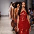 A imagem mostra um desfile da SAU Swim no SPFW N58. Uma modelo está em destaque, usando um vestido vermelho com um design intricado e recortes. O vestido tem um estilo artesanal, com uma aparência de crochê ou renda. A modelo tem cabelos longos e soltos, e usa maquiagem marcante. Atrás dela, outras modelos desfilam em fila, com roupas em tons terrosos. O público nas laterais está atento, muitos capturando o momento com seus celulares. O ambiente é iluminado, típico de um evento de moda.