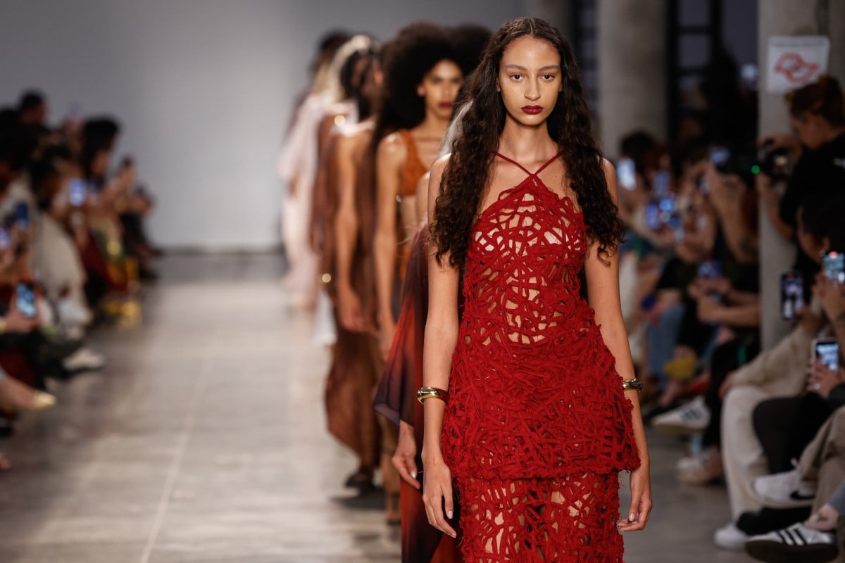 A imagem mostra um desfile da SAU Swim no SPFW N58. Uma modelo está em destaque, usando um vestido vermelho com um design intricado e recortes. O vestido tem um estilo artesanal, com uma aparência de crochê ou renda. A modelo tem cabelos longos e soltos, e usa maquiagem marcante. Atrás dela, outras modelos desfilam em fila, com roupas em tons terrosos. O público nas laterais está atento, muitos capturando o momento com seus celulares. O ambiente é iluminado, típico de um evento de moda.