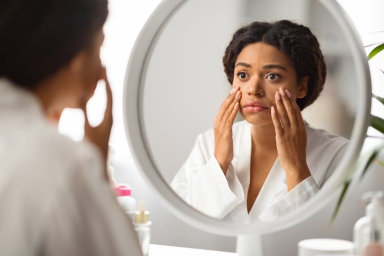 Mulher de pele negra usando robe branco, se olhando no espelho com expressão assustada e mãos no rosto 