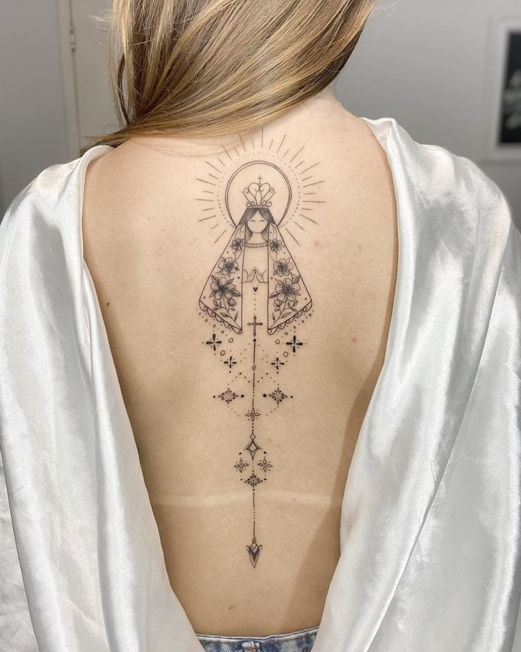 A imagem retrata uma tatuagem religiosa nas costas, representando uma figura sagrada com um manto decorado e uma coroa. O design é detalhado, com linhas finas e elementos florais, irradiando uma aura de luz. A tatuagem transmite espiritualidade e devoção, destacando-se pela elegância e simbolismo religioso profundo, capturando a essência da fé pessoal.