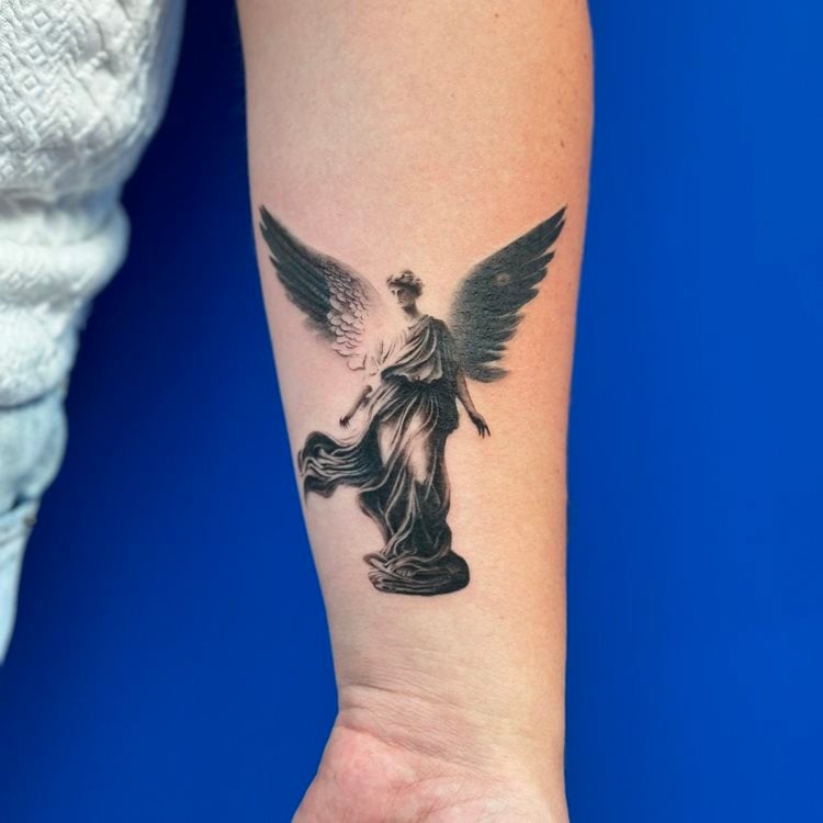 A imagem mostra uma tatuagem religiosa no antebraço, retratando um anjo com asas abertas em um fundo azul. O anjo está em uma pose graciosa, vestido com um manto esvoaçante. Os detalhes sombreados e a expressão serena da figura transmitem uma sensação de proteção e espiritualidade, simbolizando a fé e a devoção do portador.