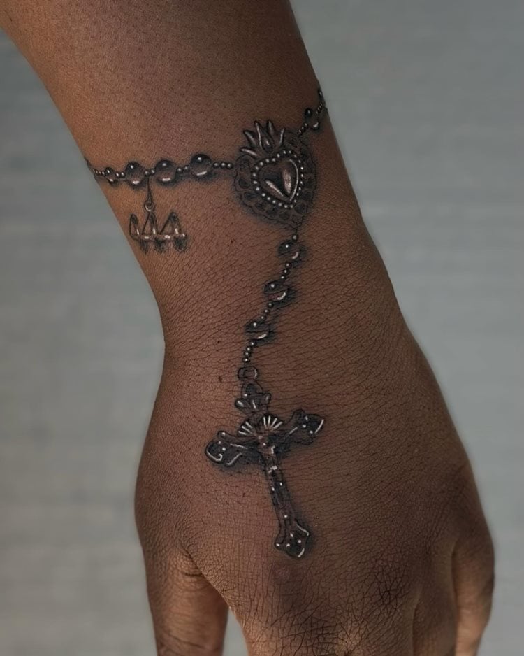 A imagem apresenta uma tatuagem religiosa no dorso da mão, representando um terço detalhado. A corrente de contas envolve o pulso, culminando em uma cruz intricada. O design inclui um coração sagrado, simbolizando devoção e fé. A arte é rica em detalhes, destacando-se pelo realismo e pela profundidade emocional que transmite ao observador.