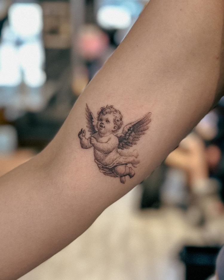 A imagem mostra uma tatuagem religiosa de um anjo no braço de uma pessoa. O anjo, delicadamente desenhado, possui asas abertas e expressão serena, simbolizando proteção e espiritualidade. Os detalhes nas asas e no corpo conferem realismo e profundidade à arte. O fundo desfocado destaca ainda mais a tatuagem, enfatizando sua importância pessoal e simbólica.