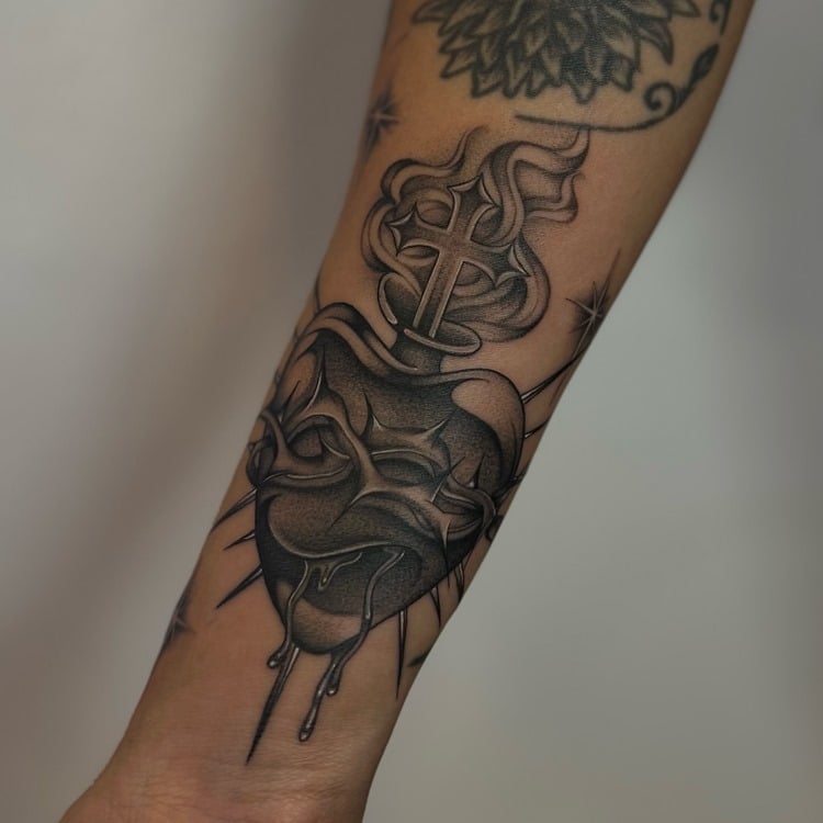 A imagem exibe uma tatuagem religiosa no antebraço, representando um coração sagrado com uma cruz e chamas. O design é detalhado, com sombreamento intricado e linhas precisas, simbolizando devoção e espiritualidade. O coração cercado por uma coroa de espinhos destaca a profundidade emocional e a fé do portador, tornando a arte um poderoso símbolo religioso.