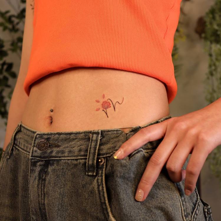 A imagem mostra a parte inferior do abdômen de uma pessoa, com uma tatuagem de flor em traços finos e pétalas rosadas. A pessoa veste um top laranja e jeans, e exibe um piercing no umbigo. A mão está levemente posicionada sobre o cós da calça, destacando o estilo casual e jovem do visual.