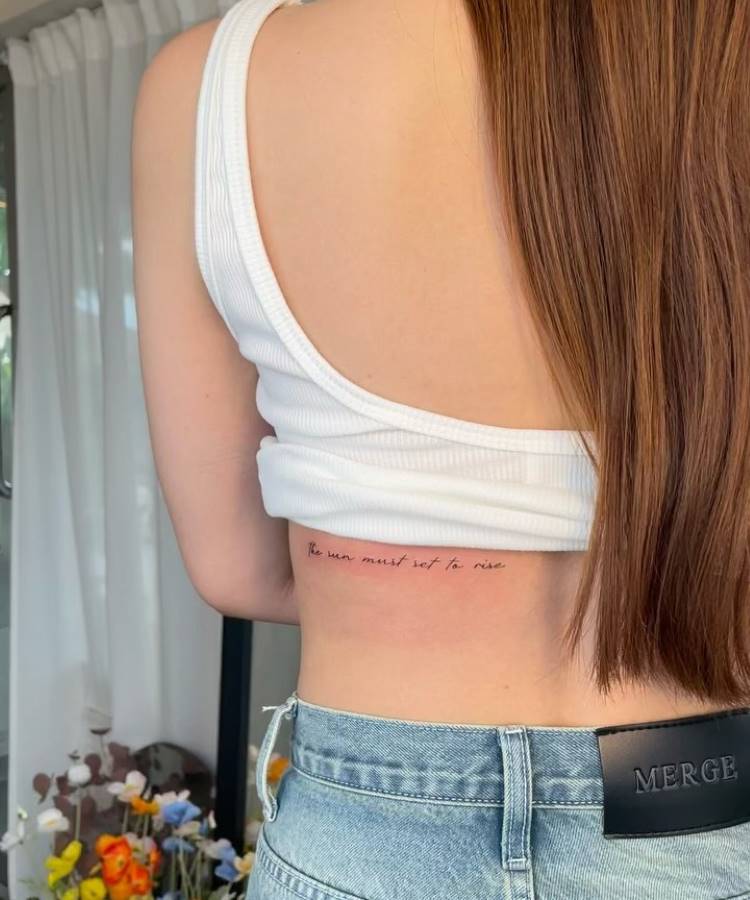 A imagem mostra uma mulher de costas, usando um top branco sem mangas e jeans claros. Ela tem cabelos longos e lisos. Na lateral do corpo, há uma tatuagem com a frase "The sun must set to rise" em letras cursivas. Ao fundo, cortinas brancas e flores coloridas complementam o ambiente iluminado.