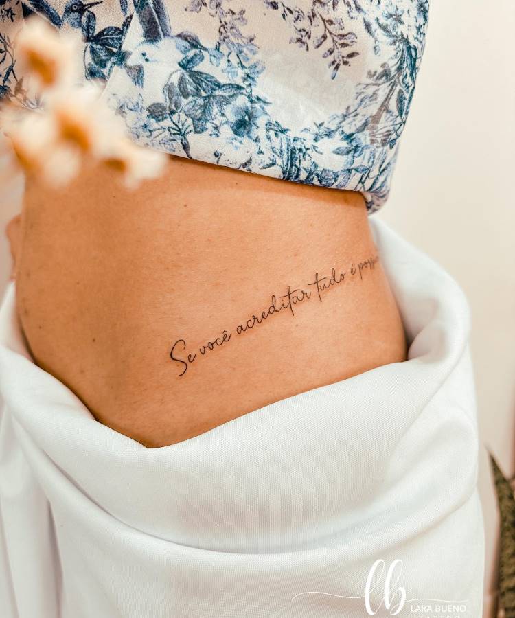 A imagem mostra uma tatuagem delicada na lateral do corpo de uma pessoa, com a frase "Se você acreditar tudo é possível." A pele está parcialmente coberta por uma blusa com estampa floral azul e branca. A cena é iluminada suavemente, destacando a simplicidade e elegância da tatuagem.