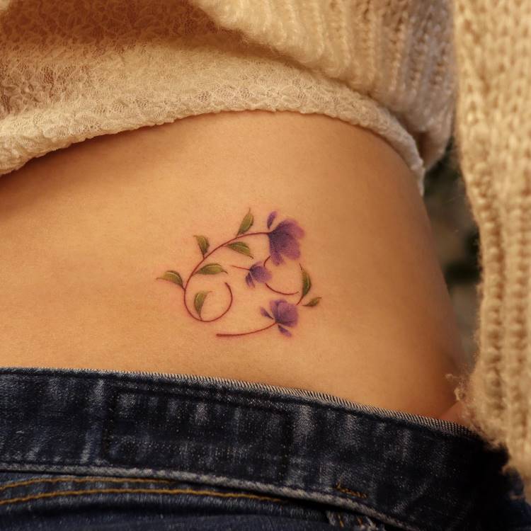 A imagem mostra uma tatuagem delicada na lateral do corpo de uma pessoa. A tatuagem é composta por flores roxas com folhas verdes, dispostas em um padrão curvo e elegante. A pele ao redor é suave, e a pessoa veste uma blusa de lã bege e calça jeans, criando um contraste harmonioso com a arte.