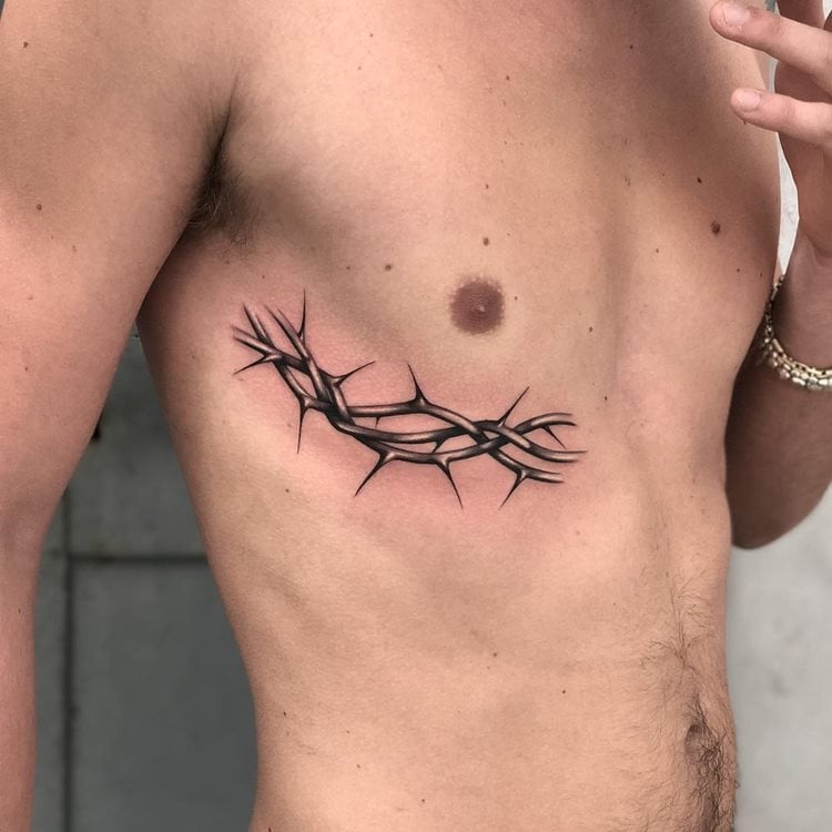 A imagem mostra uma tatuagem religiosa no lado do torso de um homem, retratando uma coroa de espinhos. Este símbolo é frequentemente associado à paixão de Cristo, representando sacrifício e fé. A tatuagem é detalhada, com sombreamento que destaca as pontas afiadas dos espinhos, criando um efeito tridimensional impressionante sobre a pele.