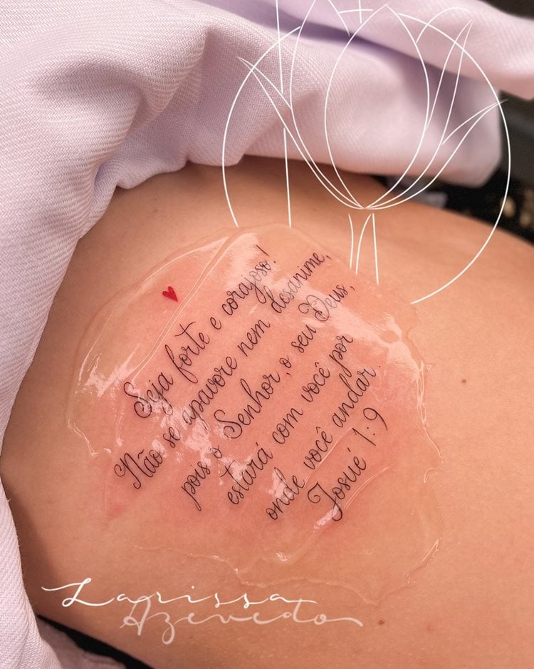 A imagem mostra uma tatuagem religiosa no braço, com o versículo de Josué 1:9: "Seja forte e corajoso!" O texto está coberto por uma película protetora, destacando a mensagem de fé e proteção divina. A tatuagem é delicada, com letras cursivas, e inclui um pequeno coração vermelho, simbolizando amor e espiritualidade.