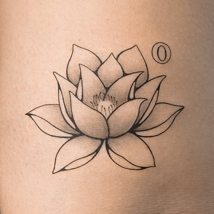 A imagem mostra uma tatuagem de flor de lótus em uma pele clara. O desenho é detalhado, com pétalas bem definidas e sombreadas, transmitindo delicadeza e equilíbrio. Ao lado da flor, há um pequeno círculo. A tatuagem simboliza pureza espiritual e renovação. O fundo é neutro, destacando a arte da tatuagem.