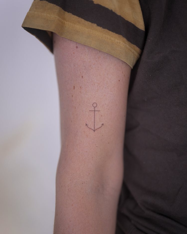 A imagem apresenta uma tatuagem de âncora no braço de uma pessoa. O desenho é minimalista e delicado, simbolizando estabilidade e esperança. A pele é clara, destacando a simplicidade do traço. A pessoa veste uma camiseta de manga curta, que deixa a tatuagem visível. O fundo é neutro, realçando a tatuagem.