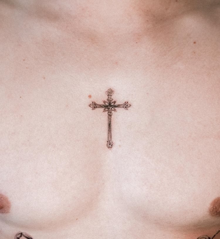 A imagem mostra uma tatuagem de cruz no centro do peito de uma pessoa. A cruz é delicada e ornamentada, destacando-se pela precisão dos detalhes. O design é simétrico e elegante, simbolizando fé e espiritualidade. A pele ao redor é clara, realçando a nitidez e a beleza do trabalho artístico.