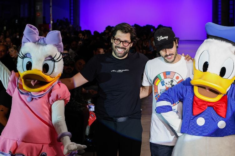 Dois homens de pele clara, um usando camiseta preta e outro usando camiseta branca com estampa colorida, abraçando pessoas duas pessoas vestidas de Margarida e Pato Donald