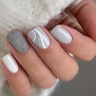 A imagem mostra uma mão com unhas bem cuidadas e pintadas em tons de branco e prata. Uma unha exibe um esmalte prateado com glitter, enquanto outra tem um design com linhas curvas prateadas e brancas sobre um fundo cinza claro. As demais unhas são pintadas de branco sólido, criando um visual elegante e sofisticado.