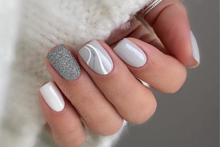 A imagem mostra uma mão com unhas bem cuidadas e pintadas em tons de branco e prata. Uma unha exibe um esmalte prateado com glitter, enquanto outra tem um design com linhas curvas prateadas e brancas sobre um fundo cinza claro. As demais unhas são pintadas de branco sólido, criando um visual elegante e sofisticado.