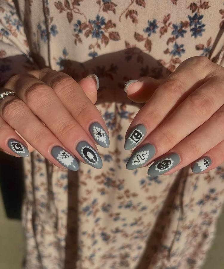 A imagem mostra unhas longas e ovais pintadas em cinza com desenhos místicos em preto e branco. Cada unha exibe símbolos diferentes, como olhos, estrelas e serpentes, criando um visual esotérico. A pessoa usa um vestido floral em tons de bege e azul, complementando o estilo boho. A iluminação destaca os detalhes artísticos das unhas.