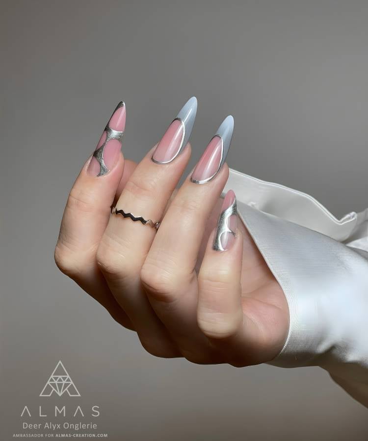 A imagem mostra unhas longas e pontiagudas com um design moderno. As unhas são pintadas em tons de cinza e rosa, com detalhes metálicos prateados em formas onduladas. O fundo é neutro, destacando as unhas. A mão está adornada com um anel fino e ondulado, e a pessoa veste uma blusa branca de cetim, criando um visual elegante.