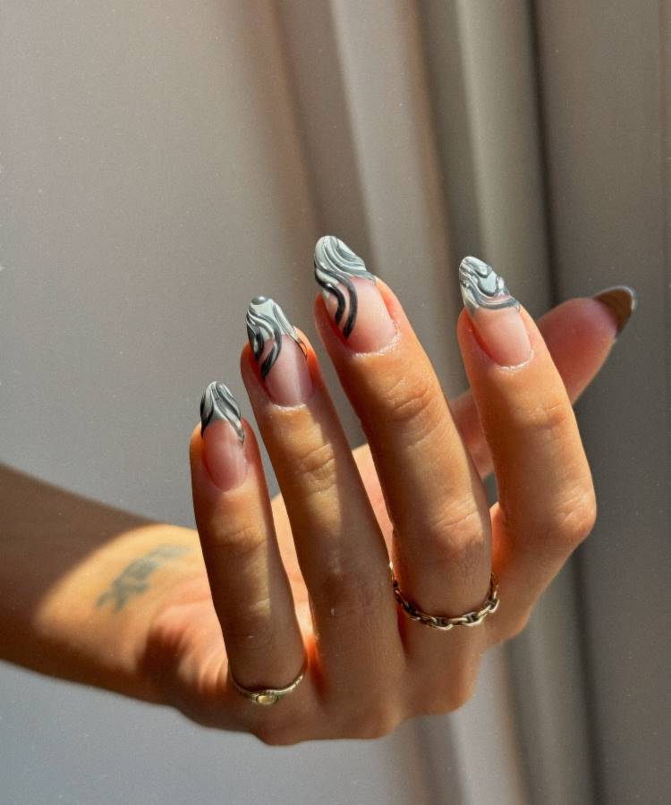 A imagem mostra unhas longas e ovais com um design elegante em tons de cinza. As pontas das unhas têm um padrão marmorizado em cinza e preto sobre uma base transparente. A mão está adornada com anéis delicados de ouro. A iluminação suave destaca o brilho e o detalhe das unhas, criando um visual moderno e sofisticado.