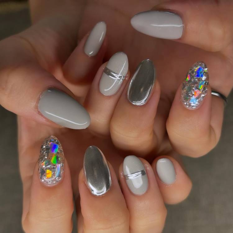 A imagem mostra unhas longas e ovais com um design sofisticado. As unhas são pintadas em tons de cinza e prateado, com algumas apresentando um acabamento metálico espelhado. Duas unhas possuem decoração com glitter holográfico, criando um efeito brilhante e colorido. O design inclui linhas prateadas horizontais em algumas unhas, adicionando um toque moderno e elegante.