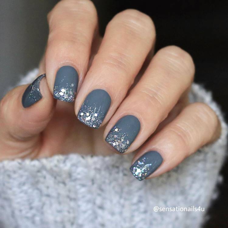 A imagem apresenta unhas pintadas, decoradas com glitter prateado nas pontas, criando um efeito degradê brilhante. As unhas são curtas e bem cuidadas. A mão está parcialmente coberta por um suéter de tricô cinza claro, que complementa o visual elegante e moderno. O design é sofisticado e atraente.