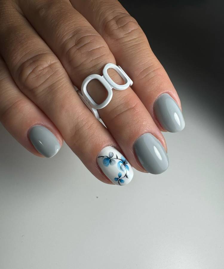 A imagem mostra unhas curtas e bem cuidadas, pintadas em um tom de cinza suave. O dedo anelar exibe um design floral delicado em azul sobre um fundo branco. A pessoa usa um anel branco estilizado, que complementa o visual moderno e elegante. O fundo é neutro, destacando a arte das unhas.