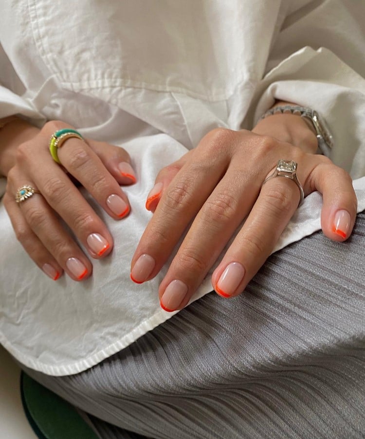 A imagem mostra mãos com unhas em estilo francesinha, com pontas laranja neon. As unhas repousam sobre um tecido branco, destacando o contraste entre o vibrante e o neutro. Anéis elegantes complementam o visual, enquanto a iluminação suave realça as cores e a textura.