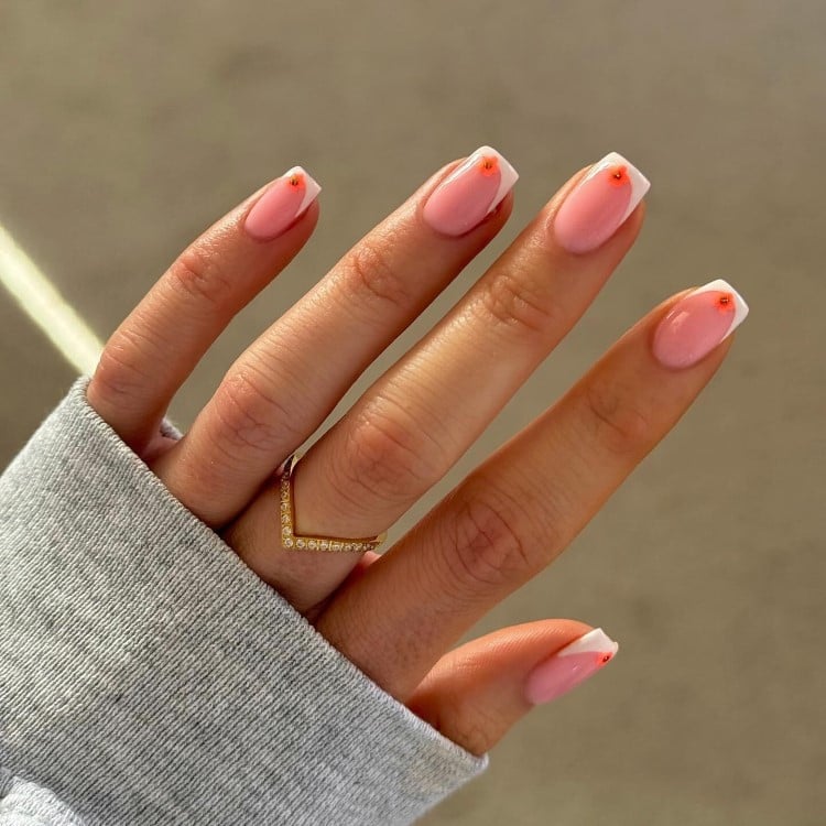 A imagem mostra uma mão com unhas em estilo francesinha, com pontas brancas e pequenos detalhes florais vermelhos. A pele suave contrasta com o fundo neutro, e um anel elegante complementa o visual. A iluminação suave destaca a delicadeza e o cuidado nos detalhes.