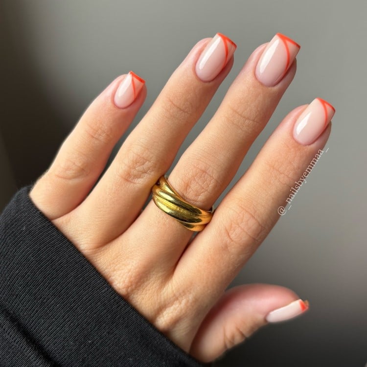 A imagem mostra unhas em estilo francesinha com pontas laranja vibrantes em formato triangular. A base é nude, destacando o contraste geométrico. As mãos usam um anel dourado sobre um fundo neutro, criando um visual elegante e moderno, com foco nas linhas ousadas.