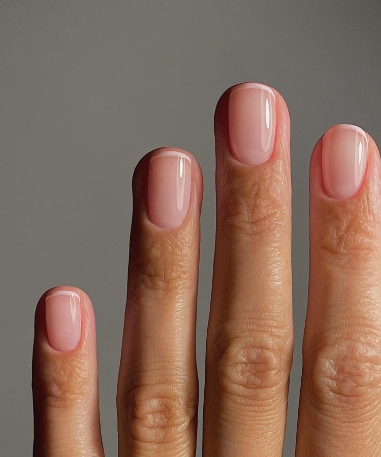 A imagem mostra uma mão com unhas em estilo francesinha, exibindo um tom suave e translúcido. O fundo neutro destaca a simplicidade e o brilho natural das unhas. A iluminação suave realça a textura e o cuidado, transmitindo uma sensação de pureza e elegância.