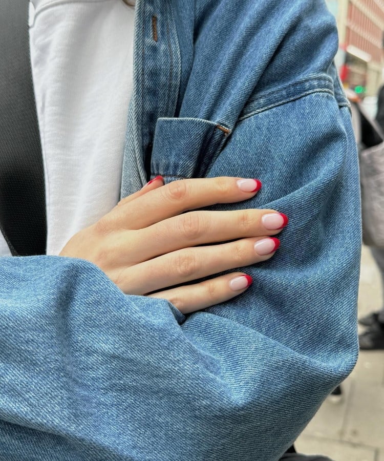 A imagem exibe mãos com unhas francesinhas de pontas vermelhas vibrantes, contrastando com o azul do casaco jeans. A pessoa usa uma camiseta branca, criando um visual casual e moderno. O fundo urbano sugere um ambiente de rua movimentada, destacando o estilo contemporâneo das unhas.