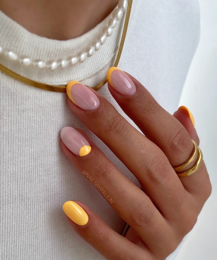 A imagem mostra unhas em estilo francesinha com pontas amarelas vibrantes sobre uma base nude. As mãos, adornadas com anéis dourados, repousam sobre um tecido branco. O contraste entre o amarelo e o nude, junto com os acessórios, cria um visual elegante e moderno.