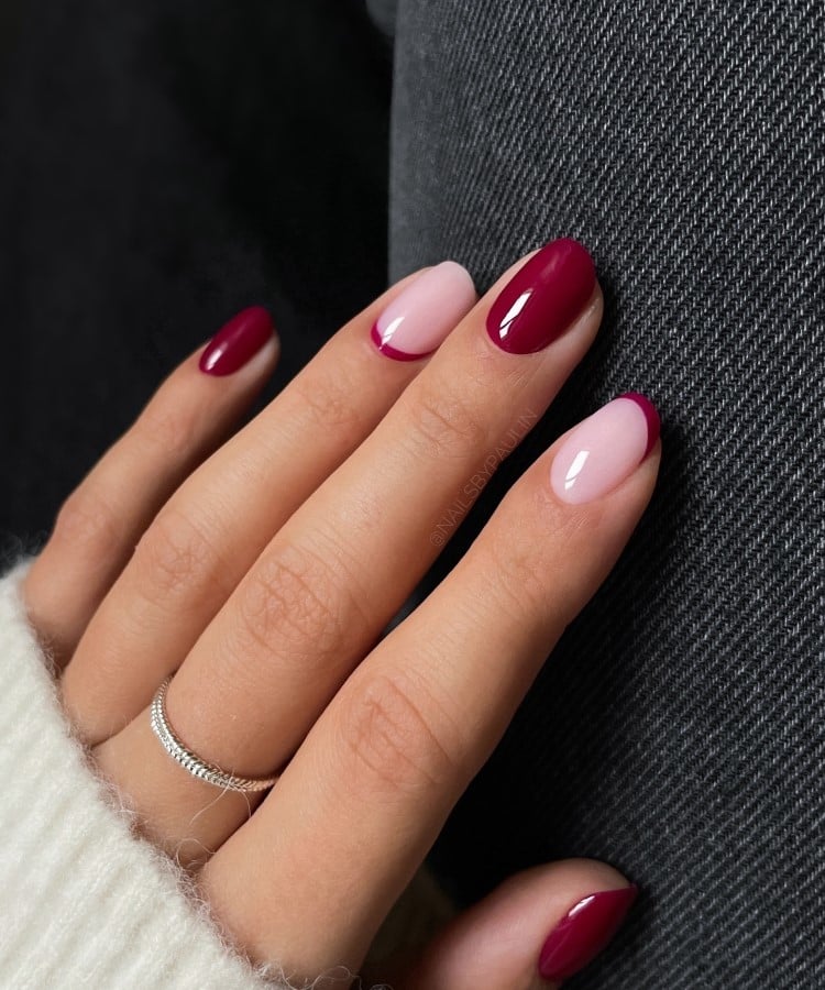 A imagem mostra unhas em estilo francesinha com pontas em tom vinho sobre uma base rosada. As unhas alternam entre francesinha e totalmente pintadas, criando um visual harmonioso. A mão repousa sobre um tecido jeans escuro, destacando o contraste entre o vinho e o fundo casual.