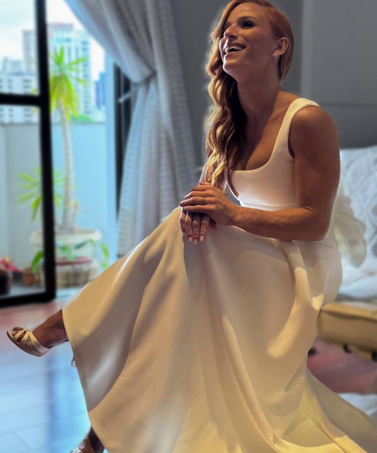 A imagem mostra Jade Barbosa de noiva no seu casamento. Ela está sentada, usando um vestido branco elegante, com um sorriso radiante no rosto. Seus cabelos ruivos caem em ondas sobre os ombros. Ao fundo, há uma janela com vista para uma área externa com plantas, e cortinas brancas que acrescentam um toque de sofisticação ao ambiente. A iluminação suave realça a beleza da noiva, capturando um momento de felicidade e serenidade antes da cerimônia.