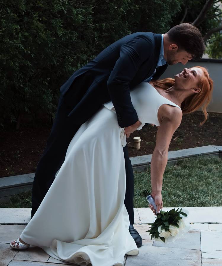 A imagem retrata os noivos Jade Barbosa e Leandro Ferlini em momento romântico em seu casamento. A mulher, com cabelos ruivos e um vestido branco elegante, está sendo inclinada para trás pelo homem, que usa um terno azul escuro. Ambos sorriem alegremente. Ela segura um buquê de flores brancas, enquanto ele a segura firmemente pela cintura. Ao fundo, há uma vegetação verde, sugerindo um ambiente externo. A cena transmite felicidade e amor, capturando um momento especial da cerimônia.