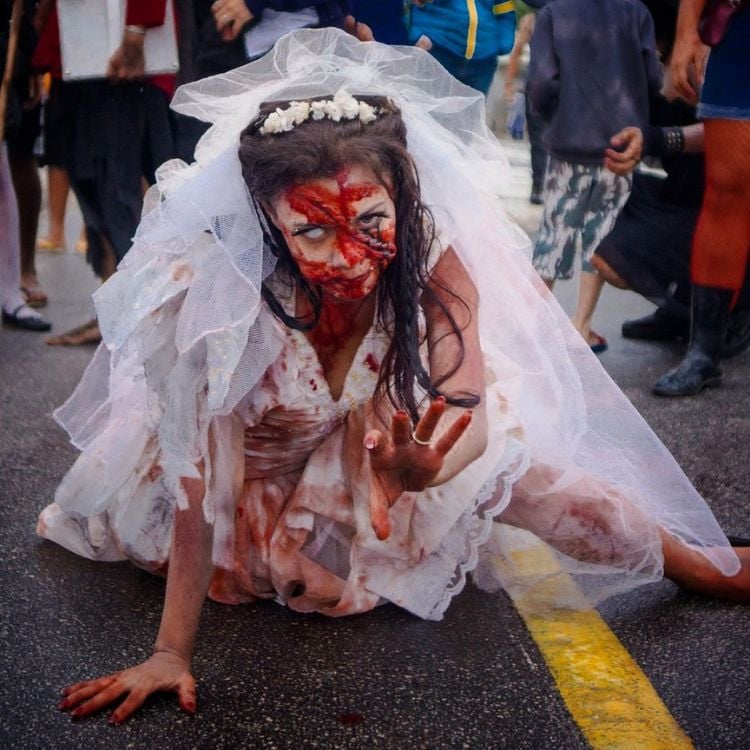 Mulher de pele clara se arrastando no chão vestida de noiva zumbi, com muito sangue falso