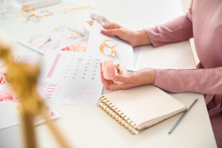 Uma pessoa, vestindo rosa, segura um porta-alianças e examina um calendário com anéis dourados estampados. A mesa está cheia de papéis de planejamento e um caderno aberto, sugerindo preparativos para um casamento ou evento especial. Tudo iluminado suavemente.