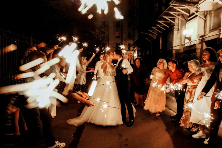 Um casal de noivos caminha à noite por uma rua, cercado por amigos e familiares segurando velas de faísca. A noiva veste um vestido branco elegante, e o noivo, um terno escuro. A atmosfera é festiva e mágica, iluminada pelas faíscas.