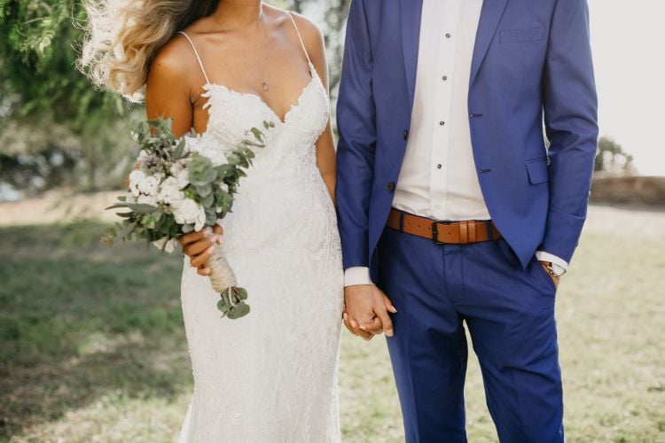 Um casal em trajes de casamento está de mãos dadas ao ar livre. A noiva veste um vestido branco rendado e segura um buquê de flores brancas e verdes. O noivo usa um terno azul com cinto marrom, em um cenário natural.