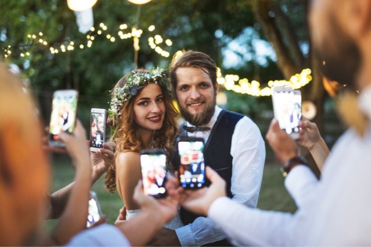 Um casal sorridente, em clima de casamento, é cercado por amigos que tiram fotos com celulares. A noiva usa uma coroa de flores e o noivo está de colete e gravata borboleta. Luzes decorativas brilham ao fundo, criando um ambiente festivo.