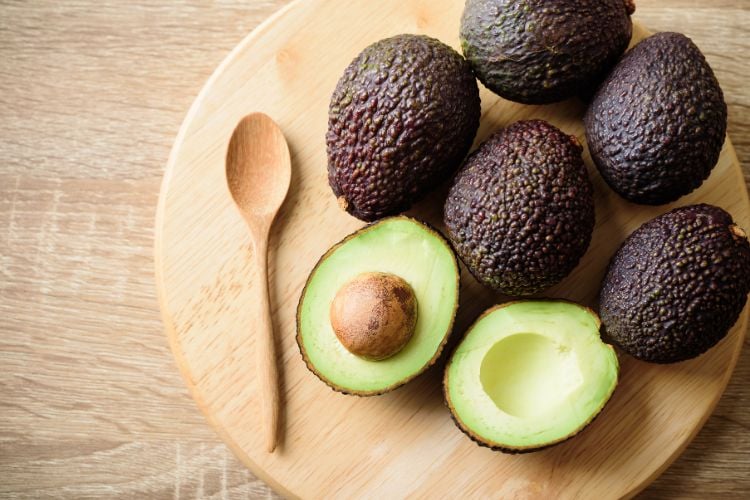 Foto de avocados abertos e cortados ao meio sobre tábua de madeira e ao lado de colher também de madeira