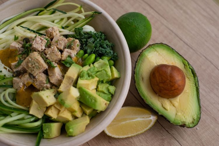 Foto de tigela com abacate, proteína e outros legumes. Ao lado, há metade de um abacate e dois limões