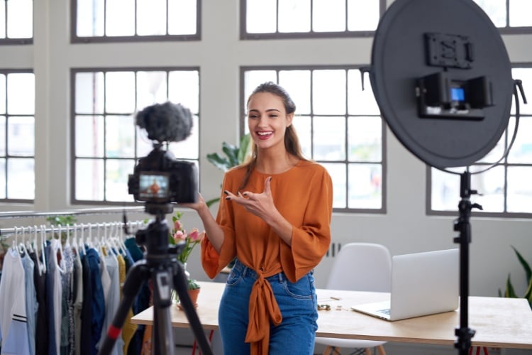 Foto de mulher de pele clara usando blusa laranja e calça jeans e gravando com câmera e microfone 