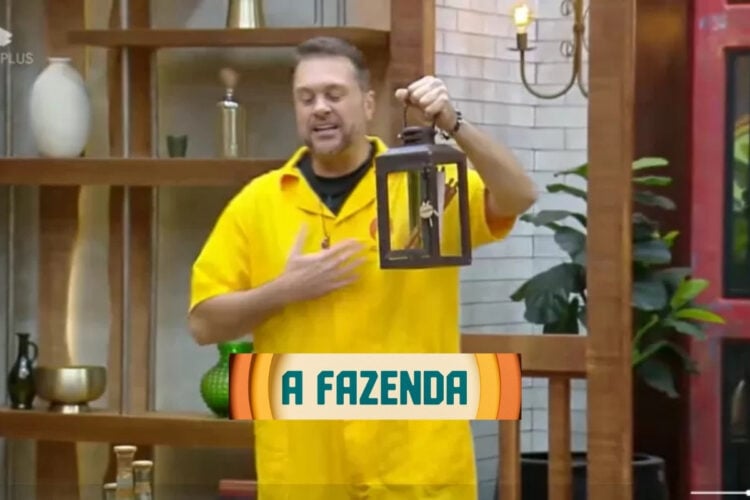 Um participante de A Fazenda 16 segura o Lampião, símbolo de poder no reality show. Ele veste uma camisa amarela e está em um cenário de pedras, expressando entusiasmo. O logotipo do programa, "A Fazenda", aparece em destaque na imagem.