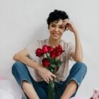 Foto de mulher de cabelos curtos e castanhos usando camiseta bege e calça jeans. Ela segura um buquê de rosas vermelhas, um dos arquétipos do amor