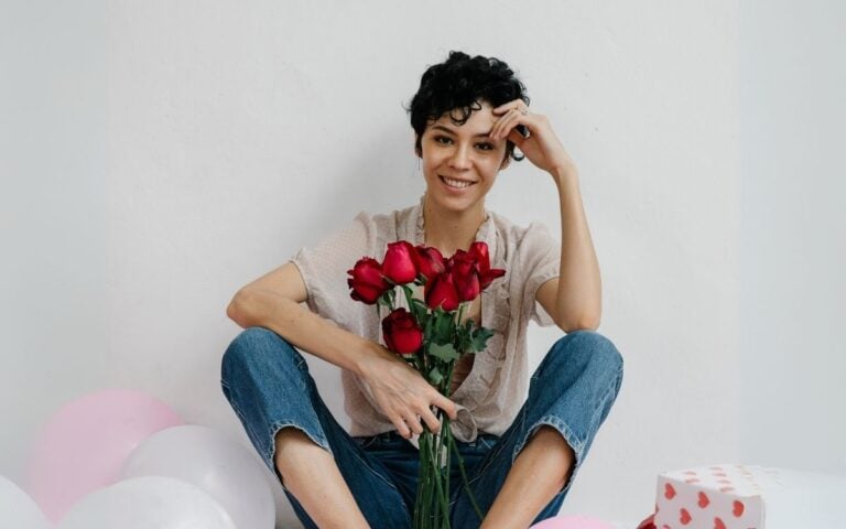 Foto de mulher de cabelos curtos e castanhos usando camiseta bege e calça jeans. Ela segura um buquê de rosas vermelhas, um dos arquétipos do amor